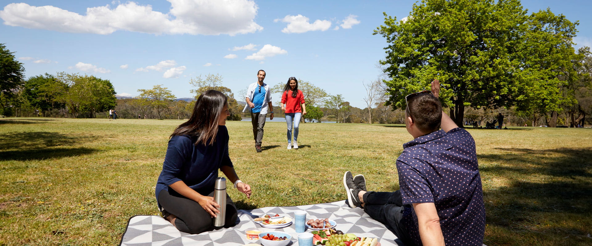 A man seated on a picnic blanket waves to two friends walking towards him on the grass as a woman kneels to set up food and drinks beside him.