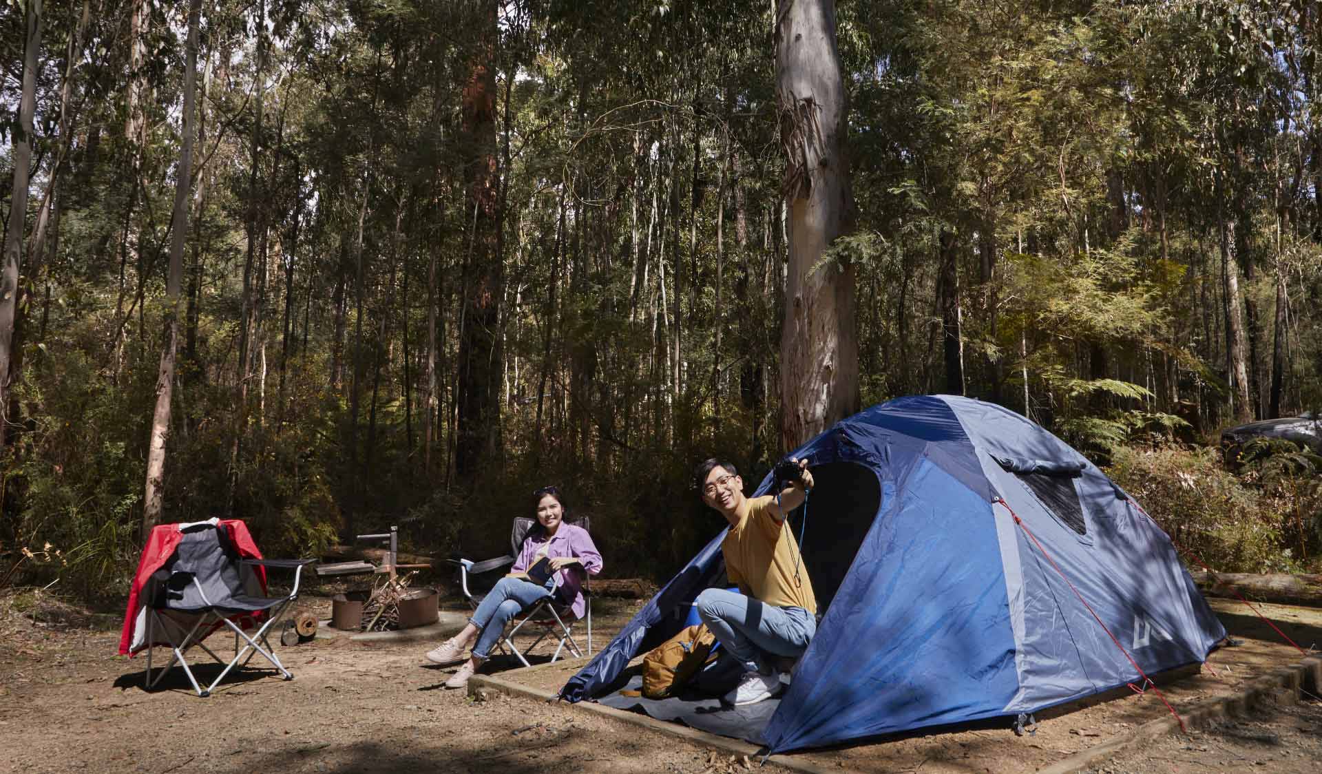 A man talks to his wife who is sitting on a camp chair as he exists his tent.