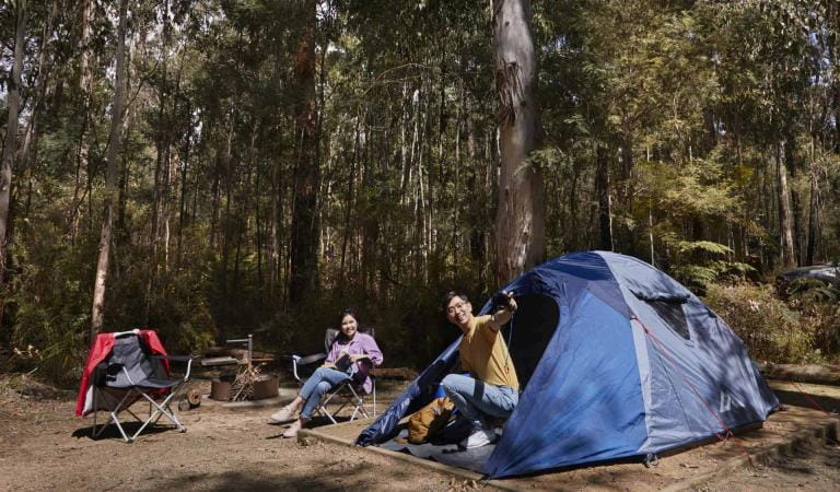 A man talks to his wife who is sitting on a camp chair as he exists his tent.