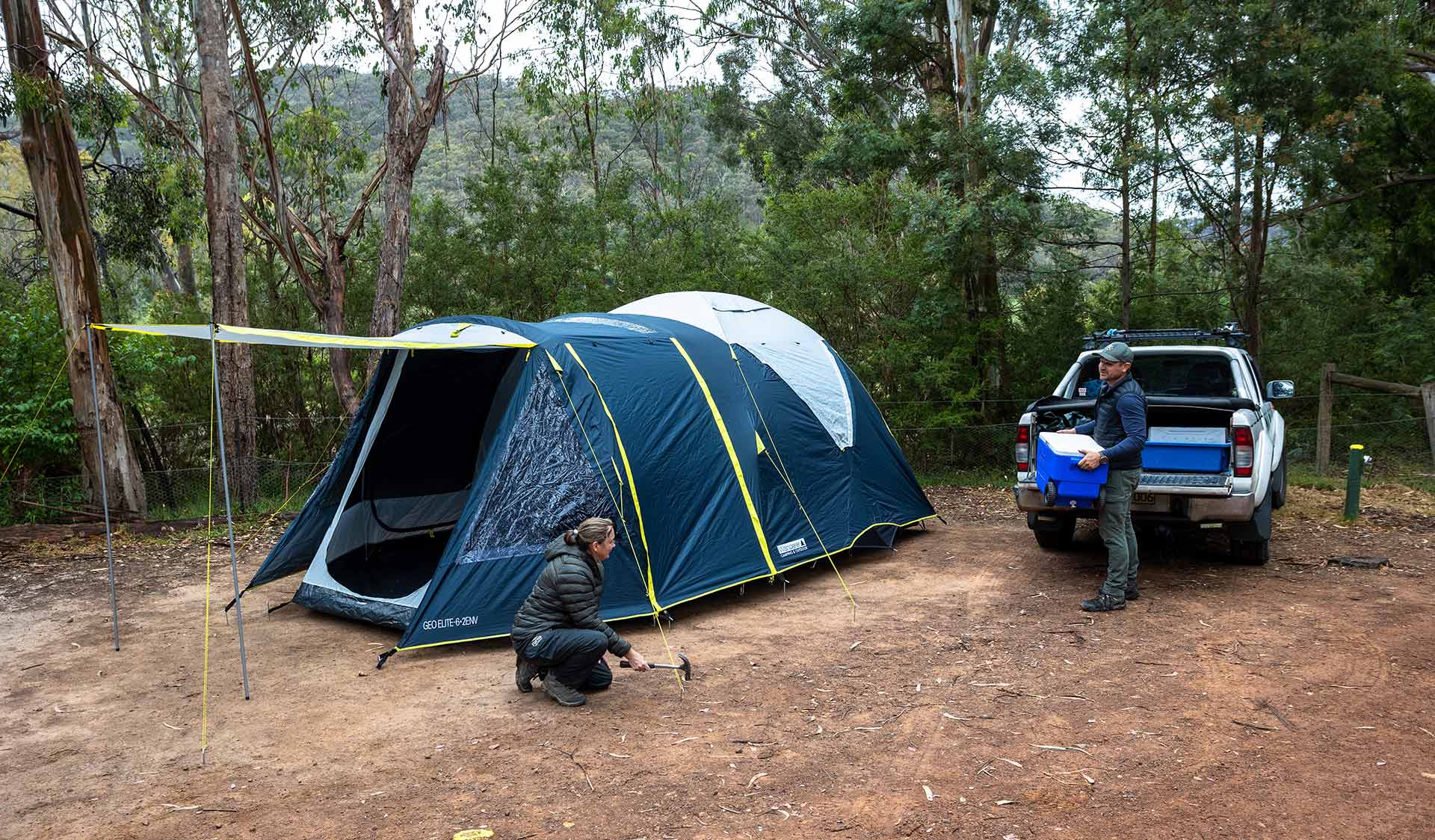 A man unpacks an esky from the tray of a ute while his partner hammers a tent peg into the ground at Candlebark Campground at Lake Eildon National Park.