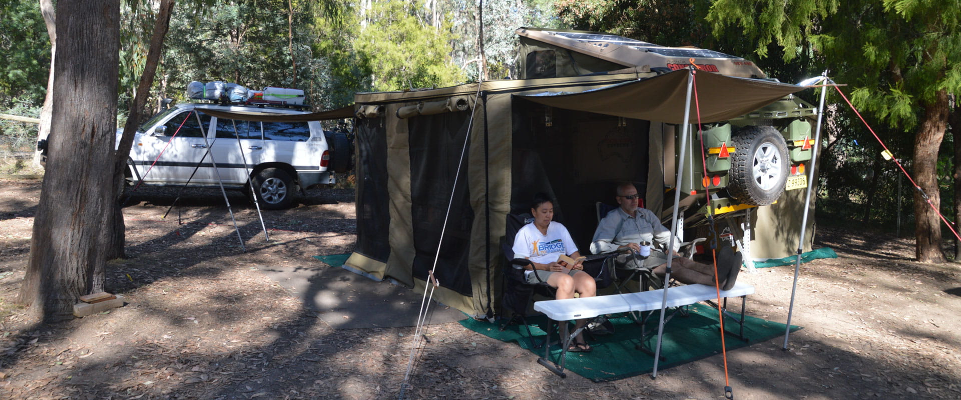 Two people sit under an awning of their tent. A car in the background. 
