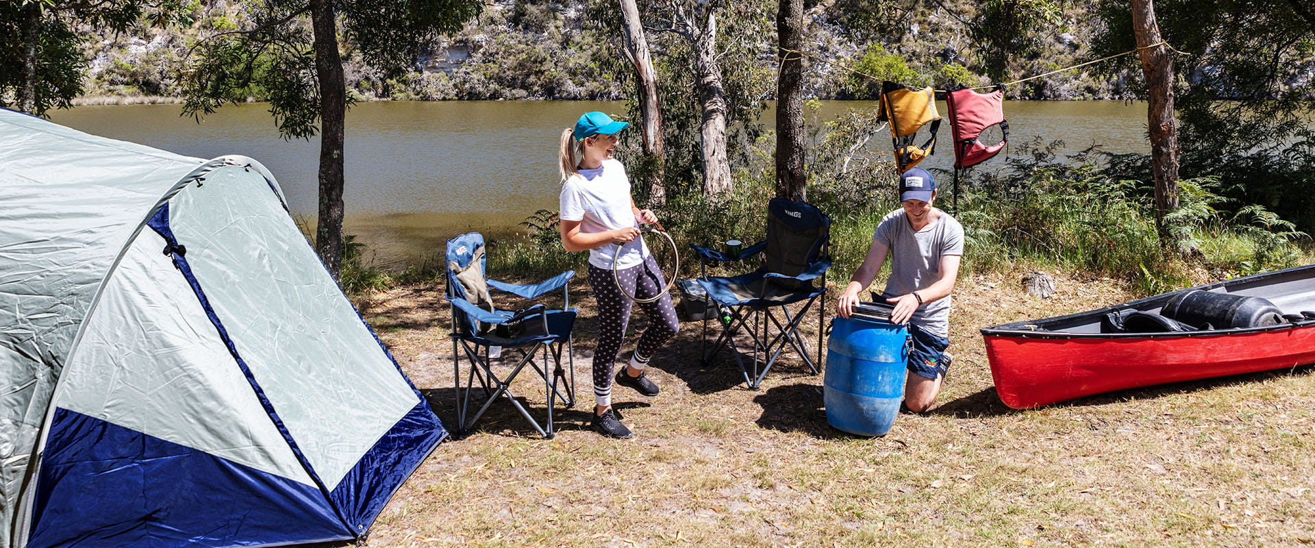 Two people unpack their kayak beside their campground. A river is in the background. 