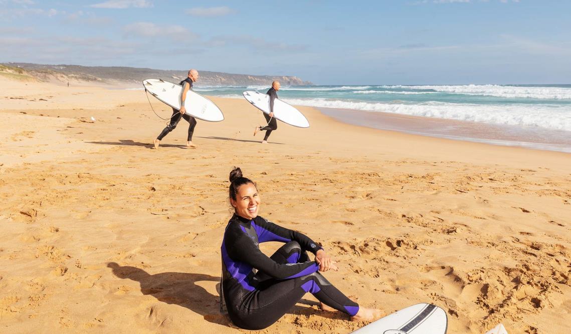 A young woman sitting on the beach in a wetsuit with two men running towards the surf at Gunnamatta Beach