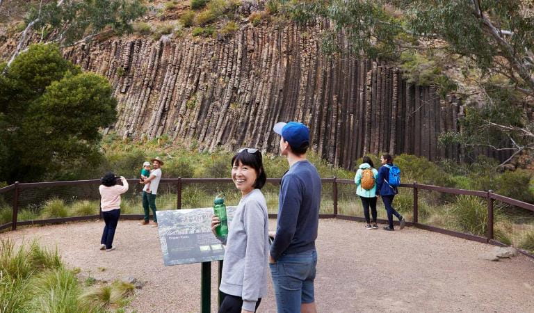 A women at the camera while others take photos of a rock formation that resembles the pipes of an organ.