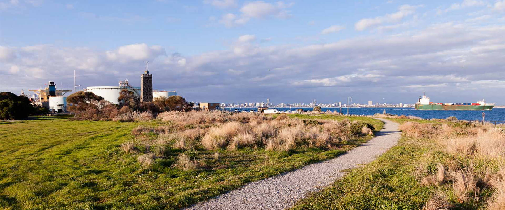 A small reserve overlooking the bay with a gravel track and native grass set amongst a green lawn, a tanker sails by in the background