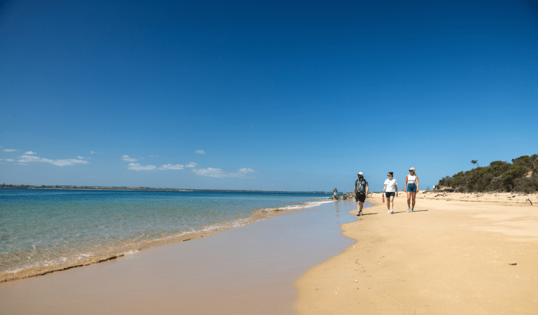 Three people walking along sand near coastline.