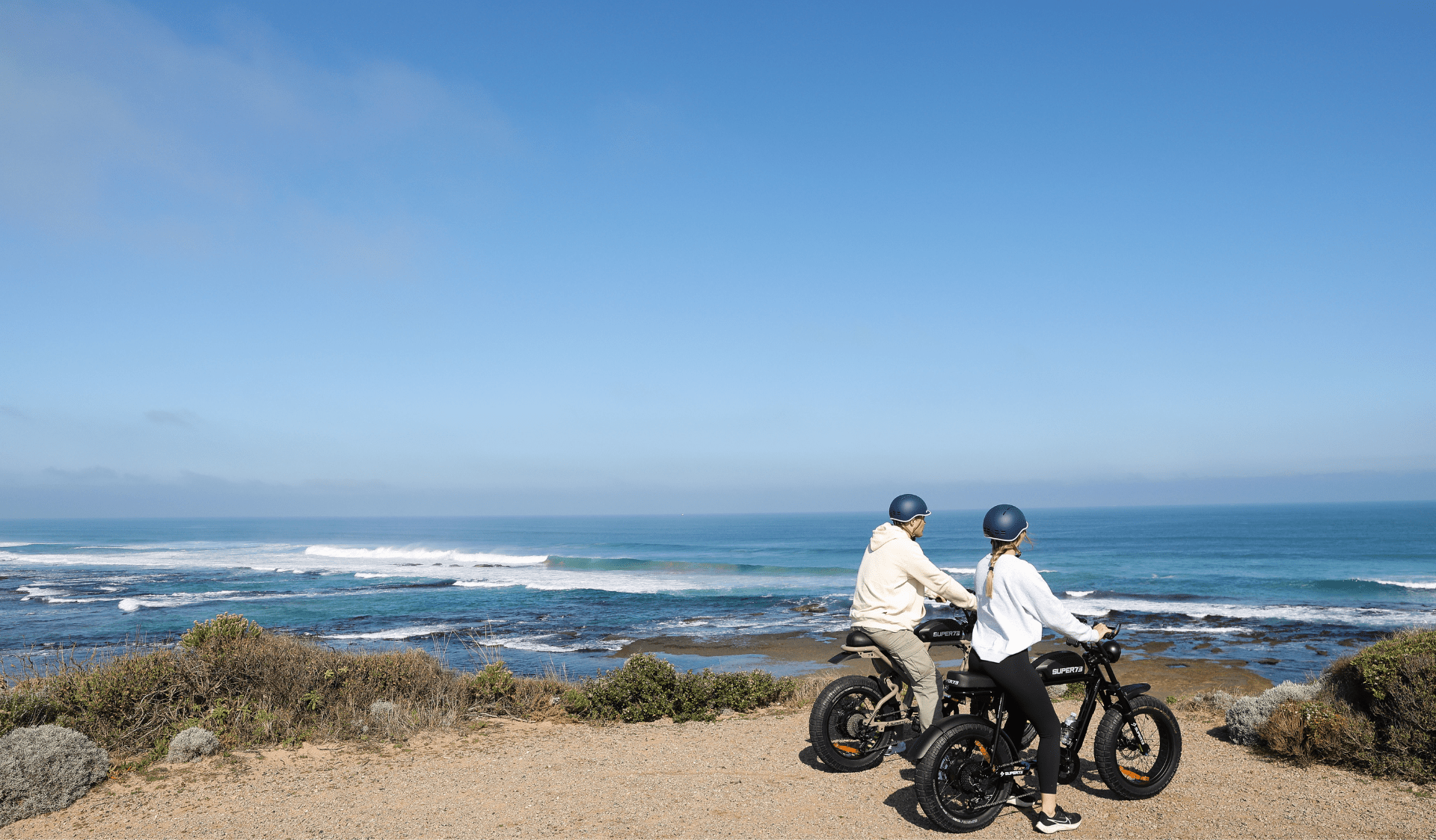Two bike riders stopped on the edge of a path looking out to the coastal view.