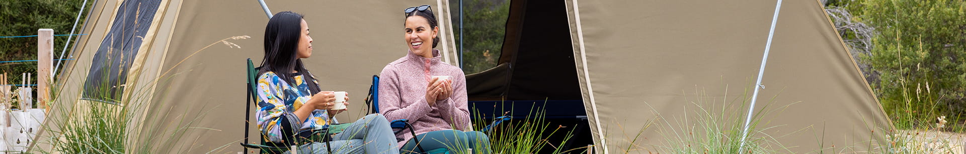 Two women smile at each other while sitting in camp chairs and holding mugs in front of a tent.