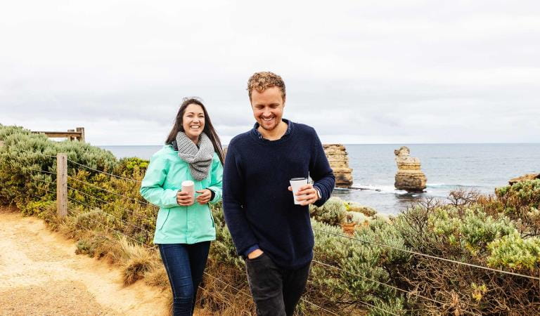 A man and woman walking with coffees at the Bay of Islands