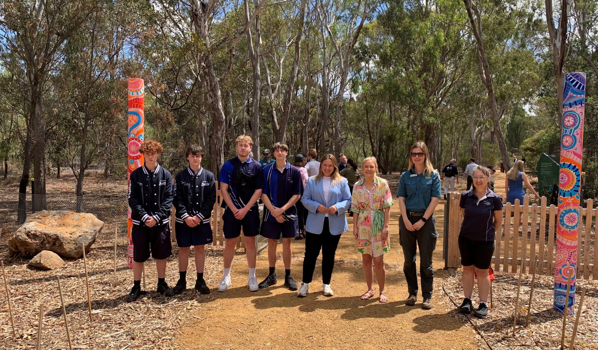 A group of people standing in front of the main entrance of the Sensory Garden. The group is standing on a gravel trail. On the right of the trail you can see a tall pole with pink, orange and blue artwork and a wooden picket fence. On the left of the trail is a tall pole with orange, red and blue artwork and a large basalt rock. The artworks include lines and symbols and were made by Wadawurrung artist Jenna Oldaker. There are tall trees in the background of the image.