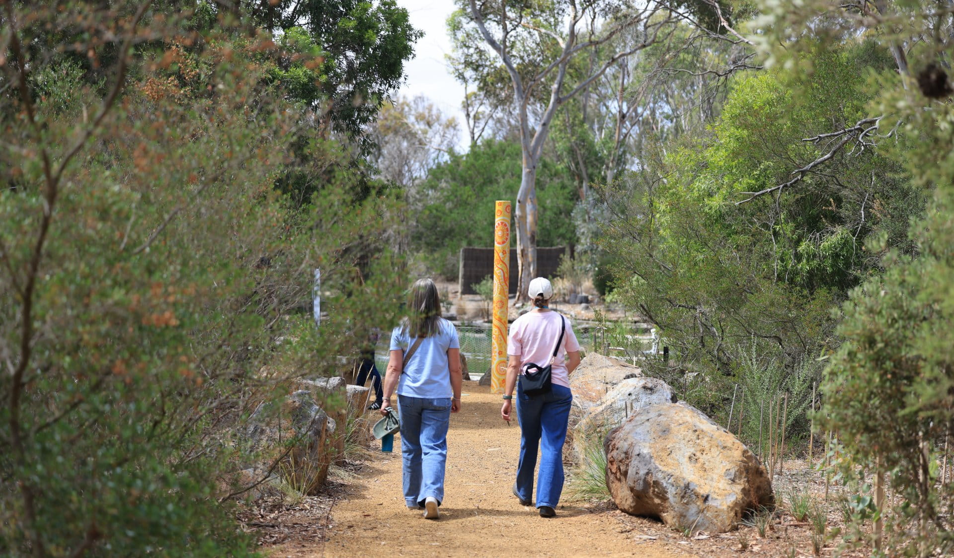 Photo of two women walking on a trail into the ‘Feel’ space in the Sensory Garden. The trail has native shrubs and basalt boulders beside it. At the back of the space you can see a tall pole with yellow and orange artwork. The artwork includes lines and symbols and was made by Wadawurrung artist Jenna Oldaker. Behind the pole you can see a pond.