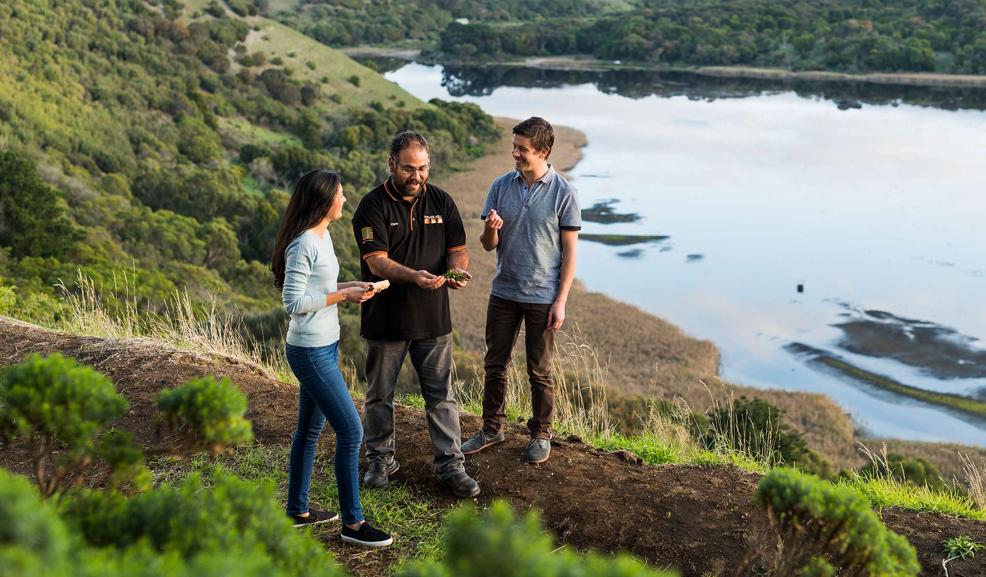 An Indigenous guide discusses traditional artifacts with a couple on a walking tour in front of the lake at Tower Hill.