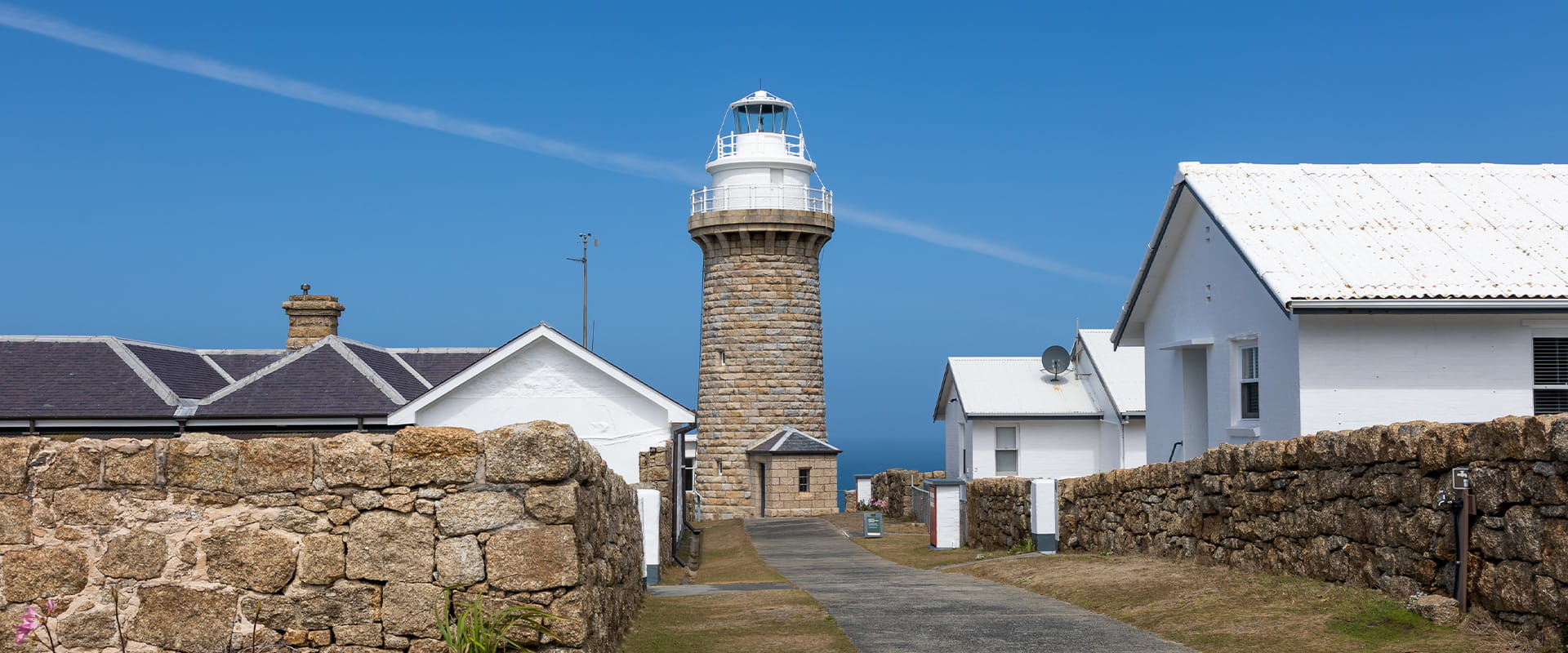 A concrete path leads to a lighthouse constructed in granite, the top of whichis painted white. Single story white buildings line the path behind a shoulder high granite wall.
