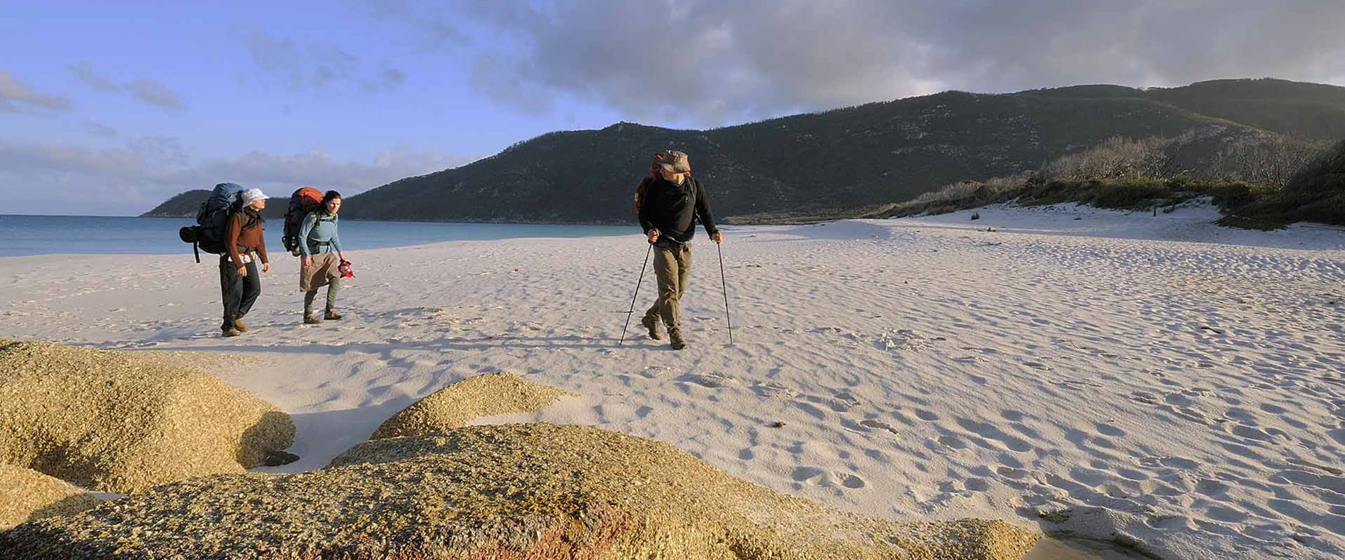 Three hikers walk across a beach with coastal mountains in the background