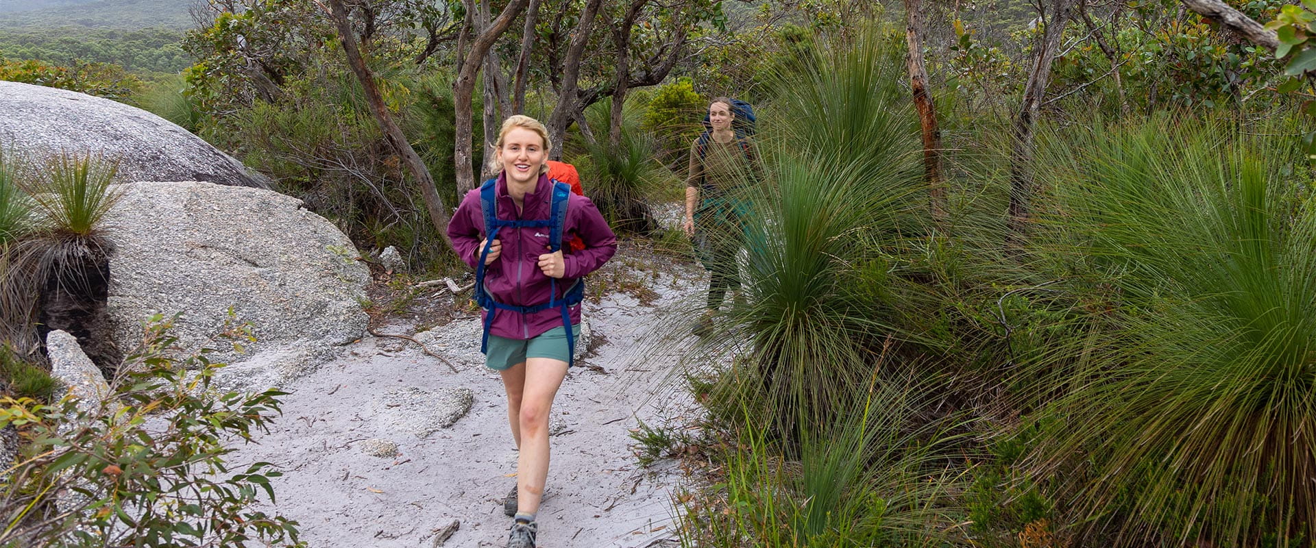 Two women in hiking gear walk along a sandy path, surrounded by boulders and vegetation.