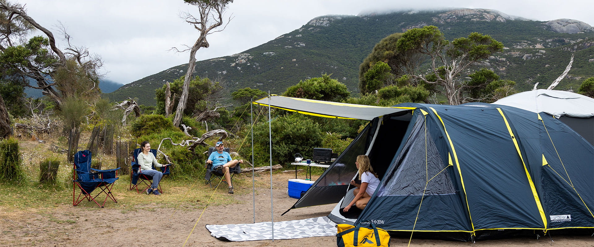 A woman and man sitting on camping chairs looking over to a woman kneeling inside an open tent.