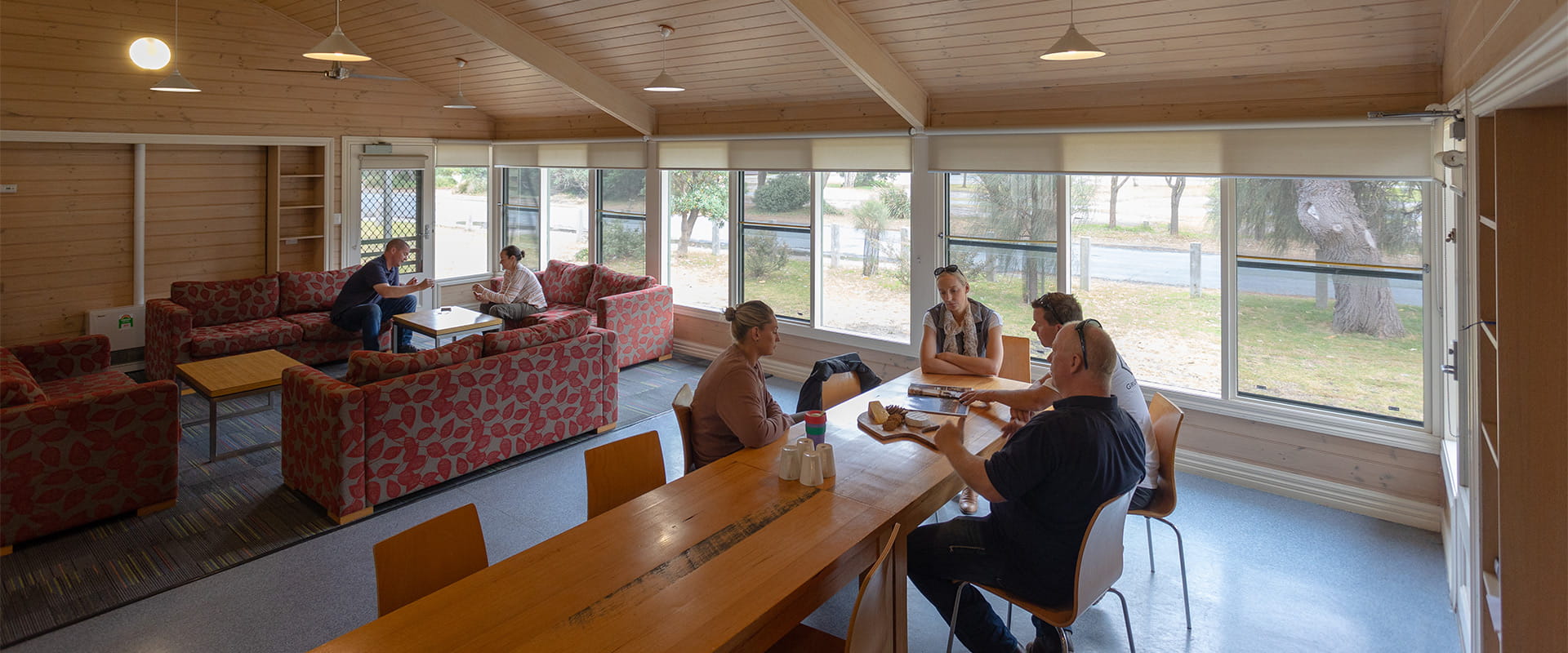 A group of four people sit at a dining table with a cheeseboard, while a man and woman play cards while sitting on sofas in the background.