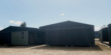 Back and front view of two cabins with a third cabin showing a built-in outdoor seating area. The cabins have green roofs, black eaves and green coloured timber cladding for the walls. There are black framed windows in the walls.