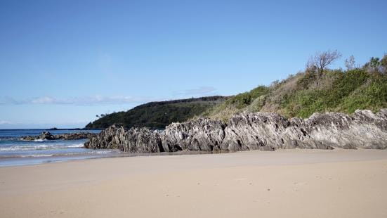 Very small waves are gently folding onto a flat almost white sandy beach. The beach shoreline has small edging of craggy rocks and grass growing on top. Above the sky is brilliant blue with a few distant puffy clouds on the horizon