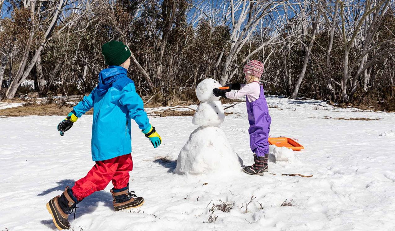 A young girl puts the final touches on her snowman as she is joined by her brother