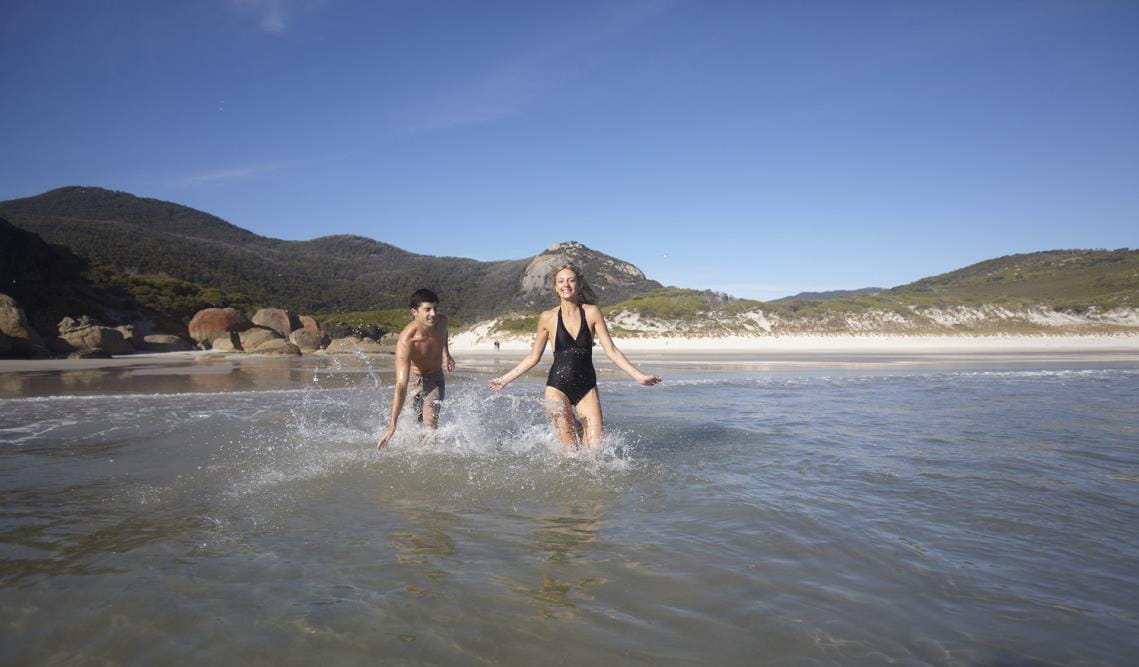 A young couple walk out into the small waves at a beach at Wilsons Promontory National Park.