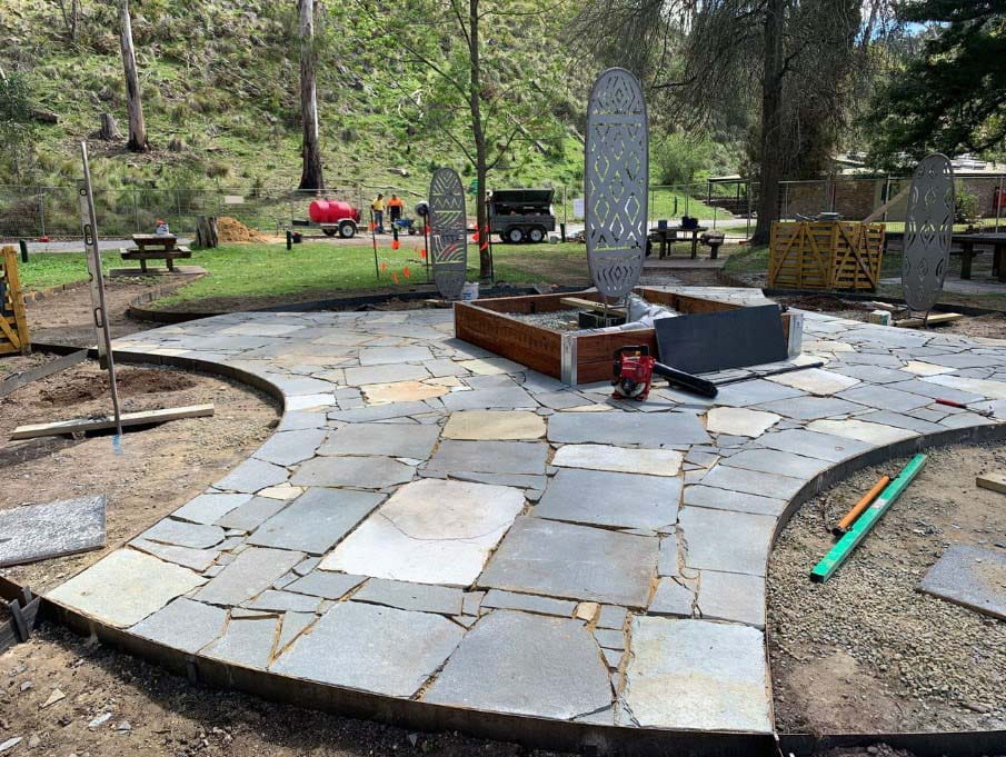 Workers stand behind bluestone and sand colour pavers which have just been laid in the Yarning Circle. There are Aboriginal symbols etched on three metal panels one in the middle and two behind. There are a green grass hill and tall trees in the background